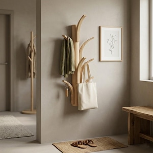May include: A wall-mounted light wood coat rack shaped like a tree with multiple branches. A green garment hangs from one branch, while a plain white canvas tote bag hangs from another. A small wooden keychain is attached to a lower branch.