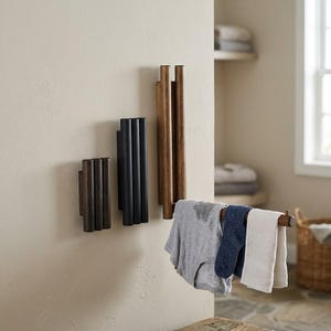 May include: Three wall-mounted wooden racks of varying heights, each featuring three vertical cylindrical dowels. The tallest rack on the right functions as a towel bar, currently holding a grey garment, a blue sock, and a white cloth.