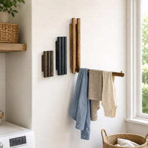 Peut inclure: Un ensemble de trois porte-manteaux muraux en bois composés de tiges cylindriques verticales. L'un des supports est déployé horizontalement pour suspendre une chemise bleue, un linge beige et un vêtement blanc à motifs.