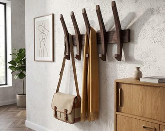 Könnte beinhalten: Eine dunkelbraune Wandgarderobe aus Holz mit fünf abgewinkelten Haken. Eine beigefarbene Umhängetasche aus Segeltuch mit braunen Lederriemen hängt am ersten Haken, und ein langer senfgelber Schal hängt am mittleren Haken der Garderobe.
