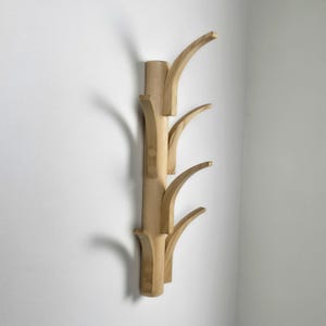 May include: A wooden wall-mounted coat rack with a natural finish. The rack features a vertical post with four curved arms extending outwards. The design is minimalist and modern, suitable for entryways or bedrooms. The rack is made of light-coloured wood.