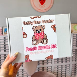 May include: A white rectangular box labeled "Teddy Bear Coaster Punch Needle Kit" with a brown teddy bear graphic and pink bow. The box is held in front of a gray wall with a pink sign that says "Our Cheery Corner".