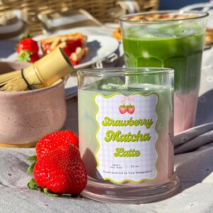 May include: A clear glass candle with a label that reads "Strawberry Matcha Latte" sits next to two fresh strawberries. The candle has a green and pink layered appearance. A glass of matcha latte is in the background.