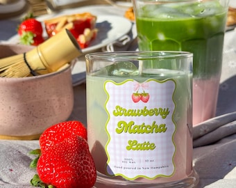 May include: A clear glass candle with a label that reads "Strawberry Matcha Latte" sits next to two fresh strawberries. The candle has a green and pink layered appearance. A glass of matcha latte is in the background.