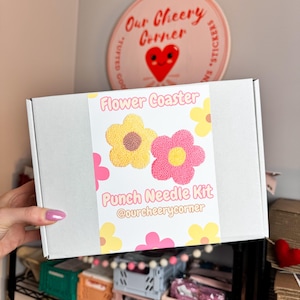 May include: A white box with a colorful label that says "Flower Coaster Punch Needle Kit @ourcheerycorner". The label features two yellow and pink flowers with brown centers.