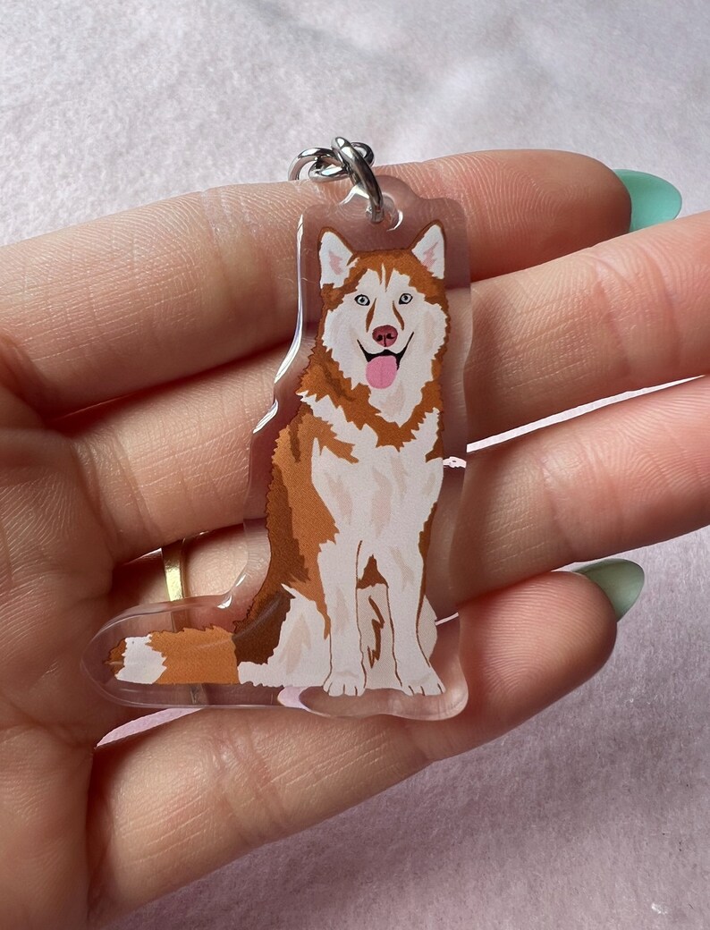 May include: A clear acrylic keychain featuring a cartoon illustration of a brown and white husky dog with a pink tongue sticking out. The dog is sitting with its paws crossed and has blue eyes.
