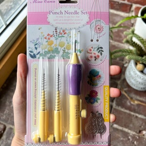 May include: A set of three punch needles for embroidery. The needles are yellow and purple with a spring mechanism. The packaging is pink and features a variety of embroidery stitches. The text on the packaging reads "Punch Needle Set" and "Quality Product".