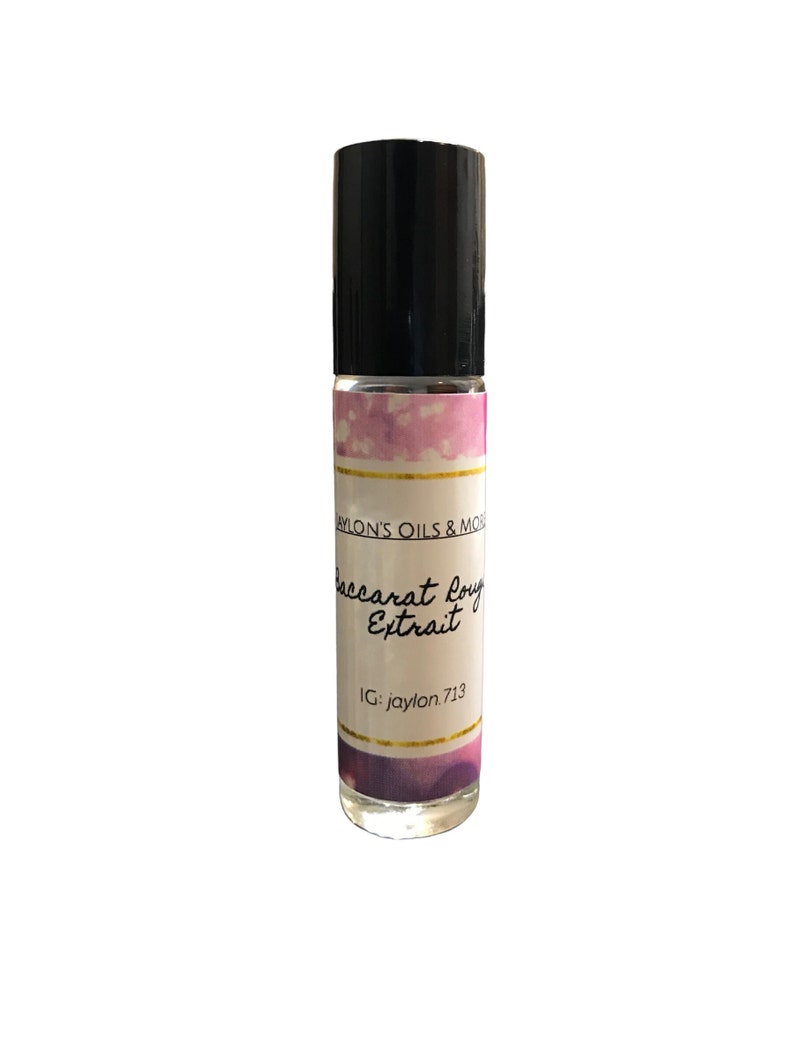 Pu&ograve; includere: Un flacone roll-on in vetro trasparente con tappo nero. L'etichetta presenta il testo "Baccarat Rouge Extrait" e "JAYLON'S OILS & MORE". L'etichetta presenta accenti rosa e viola simili ad acquerelli con bordi dorati. Viene visualizzato anche l'handle di Instagram "jaylon.713".