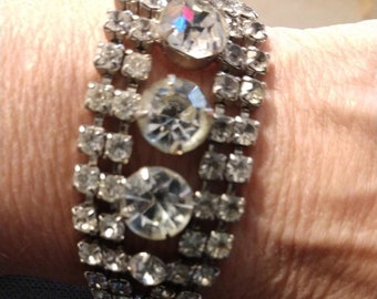 May include: A silver bracelet with multiple rows of clear rhinestones. The bracelet has a vintage style and features large, faceted rhinestones.