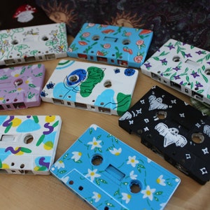 Puede incluir: Nueve cintas de cassette pintadas con diferentes diseños. Los diseños incluyen flores, formas abstractas, fantasmas y hojas. Las cintas son todas blancas con pintura de diferentes colores.