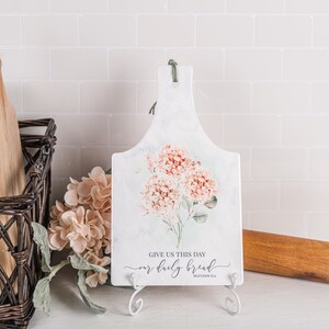 May include: White marble chopping board with a floral design of pink hydrangeas and green leaves. The board has a hanging loop and a white stand. The text "Give us this day our daily bread" is printed on the board.