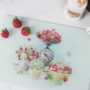 May include: A rectangular cutting board with a light blue background, featuring a vintage illustration of strawberries, flowers, and a scale. Fresh strawberries are scattered around the board. A cup of strawberries and other kitchen items are also visible.