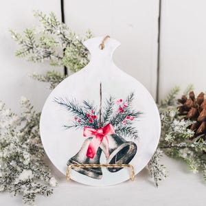 May include: White ceramic ornament shaped like a Christmas bell with a red bow and a green pine sprig with red berries. The ornament is on a gold stand.