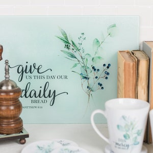 May include: A light blue glass chopping board with a watercolour design of green leaves and blue berries. The board has the text "give us this day our daily bread Matthew 6:11".