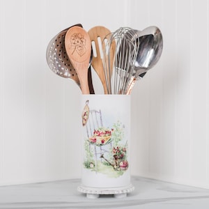 May include: A white ceramic utensil holder with a floral design, containing kitchen tools. The tools include a slotted spoon, wooden spoons, a whisk, and a ladle. The holder has a decorative base and is set against a white background.