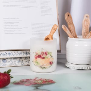 May include: A frosted glass candle holder with a floral design featuring strawberries and a wooden scoop. A white ceramic jar holds wooden spoons. A cookbook and fresh strawberries are also visible.