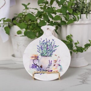 May include: White ceramic decorative plate with a lavender design. The plate features a vase of lavender and jars of lavender, with a gold-coloured metal stand. The plate has a hole at the top for hanging.