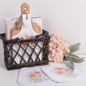 May include: A brown wicker basket with a white and pink floral design holds a stack of cookbooks, a wooden chopping board, and a white ceramic chopping board. Two white pot holders with a pink floral design and text are on the counter below the basket. A bouquet of pink hydrangeas is on the right side of the image.