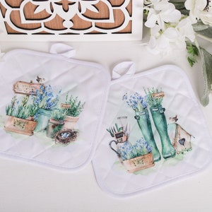 Peut inclure: Deux maniques matelassées blanches avec un motif floral. Une manique présente une pancarte qui dit "Bienvenue dans mon jardin" et l'autre présente une paire de bottes de pluie vertes.