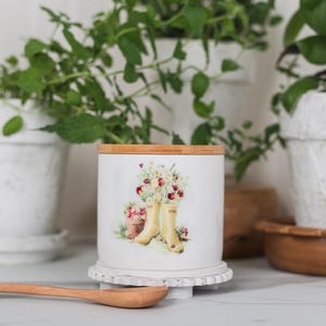 May include: White ceramic container with a wooden lid, decorated with a floral and wellington boot design. A wooden spoon is in front, with potted plants behind. The container sits on a white, decorative base.