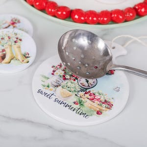 May include: Round ceramic coasters with floral designs and the text "sweet summertime." One coaster features a bicycle with flowers and fruit. A silver slotted spoon rests on one coaster. Other coasters show yellow wellington boots.