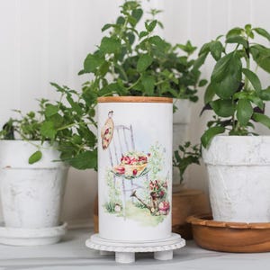 May include: A white ceramic canister with a wooden lid, decorated with a watercolour design of a chair, flowers, and fruit. The canister is on a white pedestal, surrounded by potted herbs in white pots.