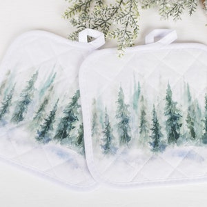 Peut inclure: Deux maniques blanches matelassées avec une scène de forêt à l'aquarelle dans les tons de vert et de bleu. Chaque manique a une boucle pour suspendre et une bordure blanche. Le motif représente des conifères.