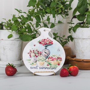 May include: A white decorative cutting board with a floral and strawberry design, and the text "sweet summertime". The board is propped up on a gold stand, with fresh strawberries in front. Potted plants are in the background.