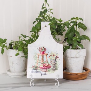 May include: A white marble-effect chopping board with a floral strawberry patch design, displayed on a white metal stand. The board features a watercolour illustration of strawberries and flowers. Potted plants surround the board.