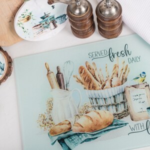 May include: A light blue glass chopping board with a watercolour illustration of bread, a jug, a rolling pin, a whisk, and a bluebird. The text "SERVED fresh DAILY" and "WITH love" is printed on the board.