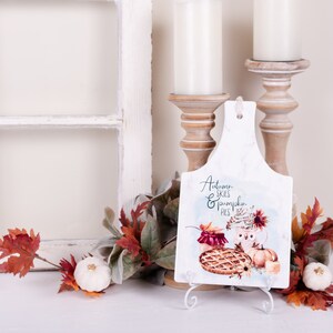 Peut inclure: Planche à découper en céramique blanche avec un motif aquarelle d'une tarte à la citrouille, un pot de confiture et une tasse de café. La planche porte l'inscription "Autumn Skies & Pumpkin Pies". La planche est sur un support blanc et est entourée de feuillage d'automne.