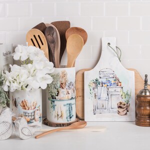 May include: A white ceramic utensil holder with a country kitchen design, featuring a wooden spoon, a wooden spatula, and a wooden ladle. The holder is sitting on a wooden base. There are two wooden salt and pepper shakers with silver tops, a white chopping board with a country kitchen design, and a white ceramic vase with white flowers.