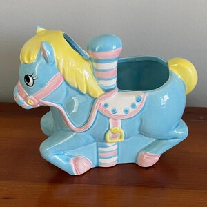 May include: A blue ceramic carousel horse planter with pink and yellow accents. The horse has a saddle and a striped pole.