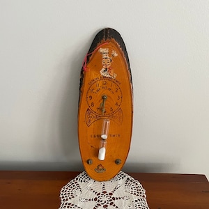 May include: A wooden egg timer with a clock face and a sand timer. The clock face has the words "Cooking & Baking Timer" and "Take Out At". The sand timer is labeled "Egg Timer".