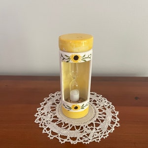 May include: A yellow and white ceramic hourglass with a sunflower design. The hourglass is sitting on a white doily.