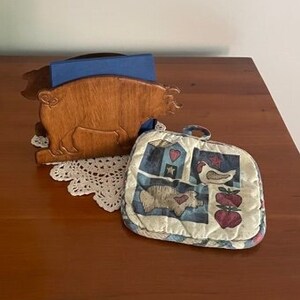 May include: Wooden pig napkin holder with a blue napkin inside, sitting on a wooden table next to a quilted pot holder with a farmhouse design featuring a house, a chicken, and apples.