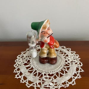 May include: A small ceramic figurine of a gnome wearing a green hat and holding a small hourglass. The gnome is painted in shades of brown, red, and white. The figurine is sitting on a white doily.