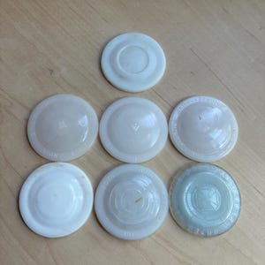 May include: Seven circular white glass jar lids arranged in a pyramid formation. Each lid features a raised center and embossed text around the rim, with some lids appearing opaque white and one appearing translucent with a visible cross emblem.