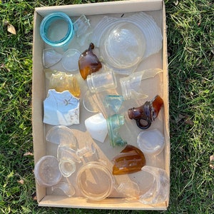 May include: A cardboard box filled with various pieces of broken glass, including clear, blue, brown, and white glass. The glass pieces are arranged in a random pattern.