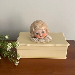 May include: A vintage cream-colored trinket box with a porcelain doll head on the lid. The doll has blonde hair, blue eyes, and a pink and white lace collar.