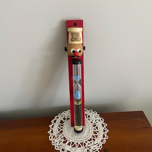 May include: A red wooden hourglass with a cartoon chef face at the top. The chef is wearing a white hat with the words "The Chef" on it. The hourglass is filled with blue sand.