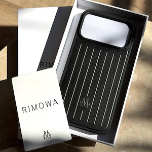 Pode incluir: Uma capa de smartphone preta com um padrão de ranhuras verticais e um pequeno logótipo, apresentada numa caixa branca. Um cartão branco com o texto RIMOWA e um logótipo está colocado à frente da caixa. A capa tem um recorte retangular grande.