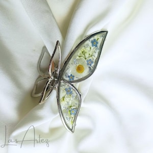 May include: A butterfly-shaped hair claw clip with a silver-toned metal frame. The wings are filled with clear resin containing pressed white daisies, small blue forget-me-not flowers, and scattered silver leaf flakes for a decorative finish.