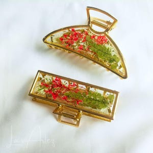 Könnte beinhalten: Zwei goldfarbene Haarspangen mit transparenten Harzrahmen, in die gepresste rote Blumen und grünes Blattwerk eingebettet sind. Eine Spange ist eine halbkreisförmige Krallenklammer, die andere eine rechteckige Spange. Beide haben einen Metallfeder.