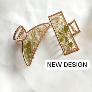 Pu&ograve; includere: Due fermagli per capelli dorati con fiori pressati. I fermagli sono di forma rettangolare e a mezzaluna. Il testo "NEW DESIGN" &egrave; stampato sotto i fermagli.