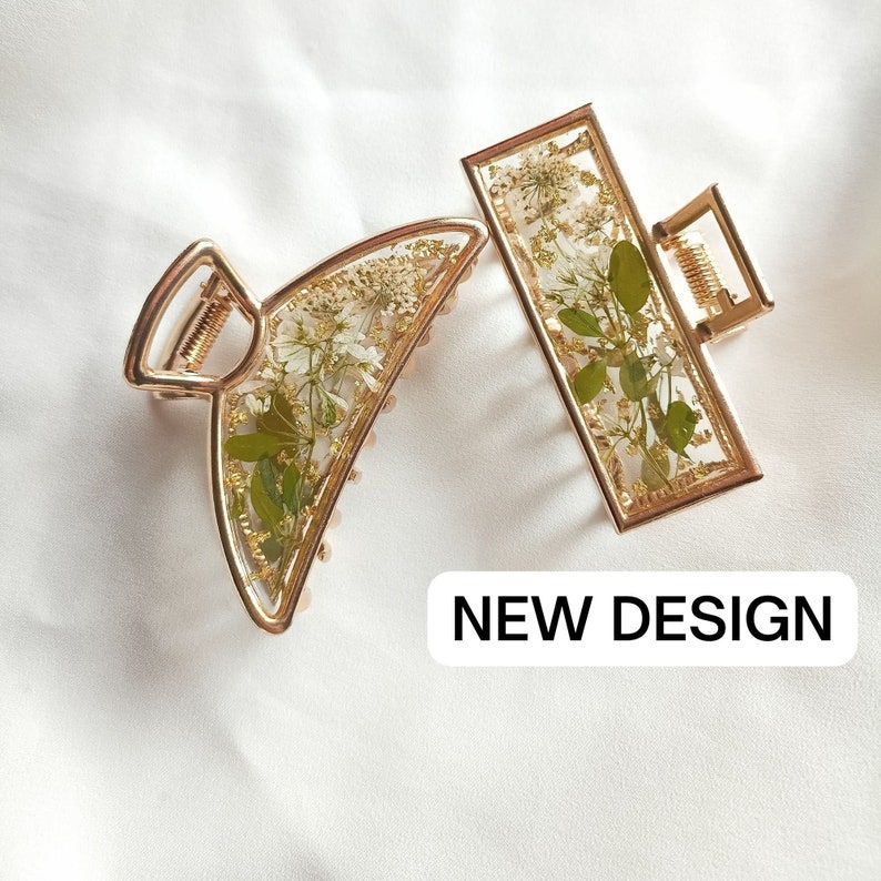 Pu&ograve; includere: Due fermagli per capelli dorati con fiori pressati. I fermagli sono di forma rettangolare e a mezzaluna. Il testo "NEW DESIGN" &egrave; stampato sotto i fermagli.
