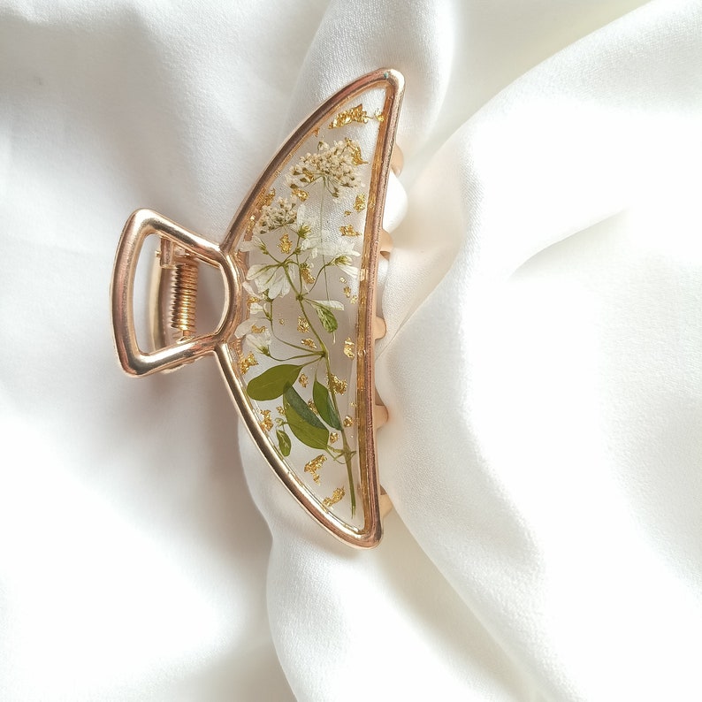 Pu&ograve; includere: Fermaglio per capelli dorato a forma di mezzaluna con un design in resina trasparente con fiori bianchi e foglie verdi.