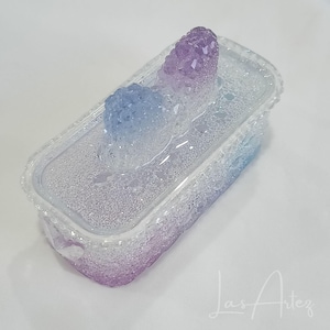 May include: A rectangular, clear container with a textured surface and a lid, featuring two crystal formations. The crystals transition in colour from purple to blue. The container is a gradient of purple and blue.