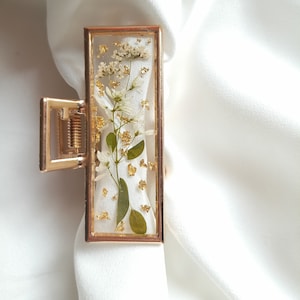 Pu&ograve; includere: Fermaglio per capelli dorato con una cornice rettangolare trasparente che contiene fiori bianchi pressati e foglia d'oro.