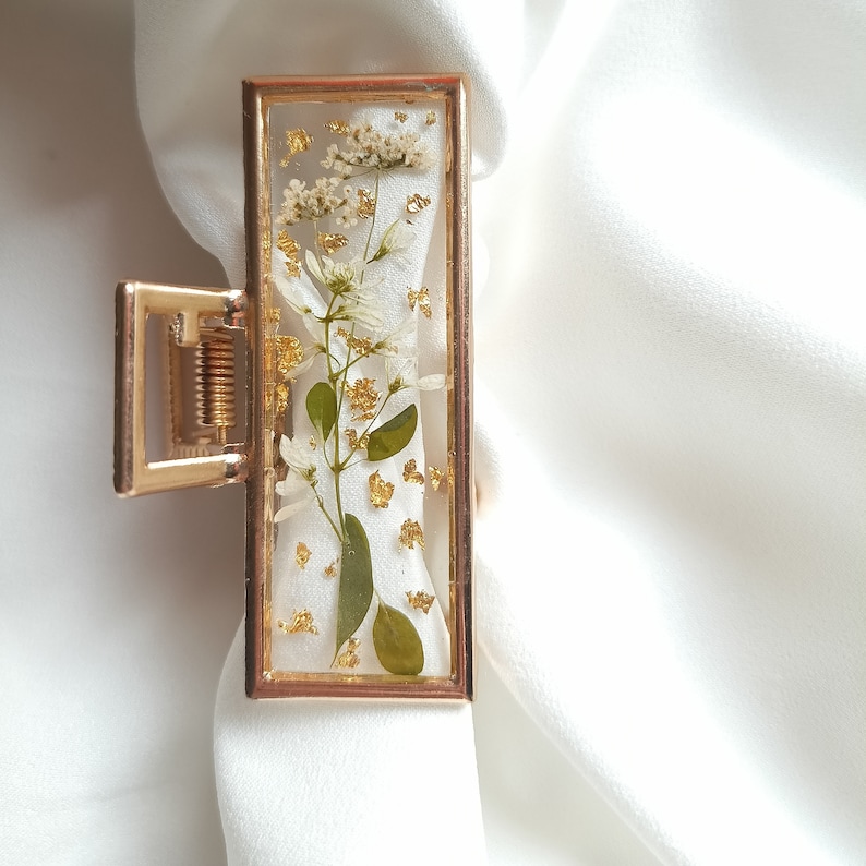 Pu&ograve; includere: Fermaglio per capelli dorato con una cornice rettangolare trasparente che contiene fiori bianchi pressati e foglia d'oro.